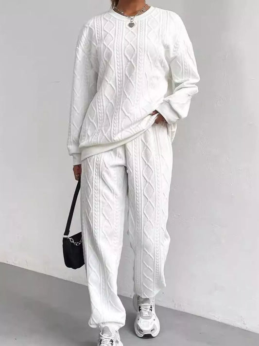 Person wearing a white cable knit sweater and pants with sneakers, holding a black bag against a plain background.