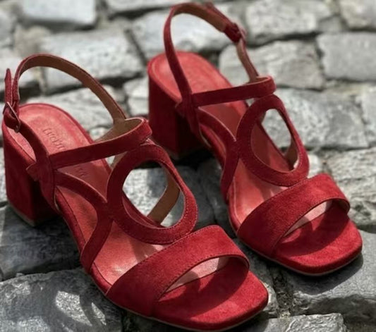 A pair of red suede block-heel sandals with crisscross ankle straps, displayed on a gray stone surface.