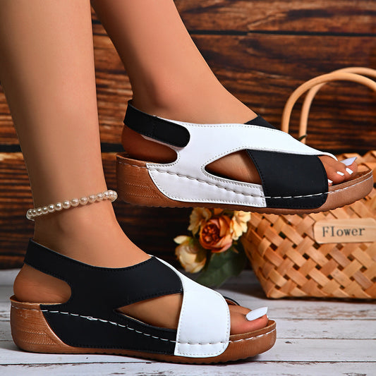 Black and white sandals with brown soles worn by a person, with a wooden background.