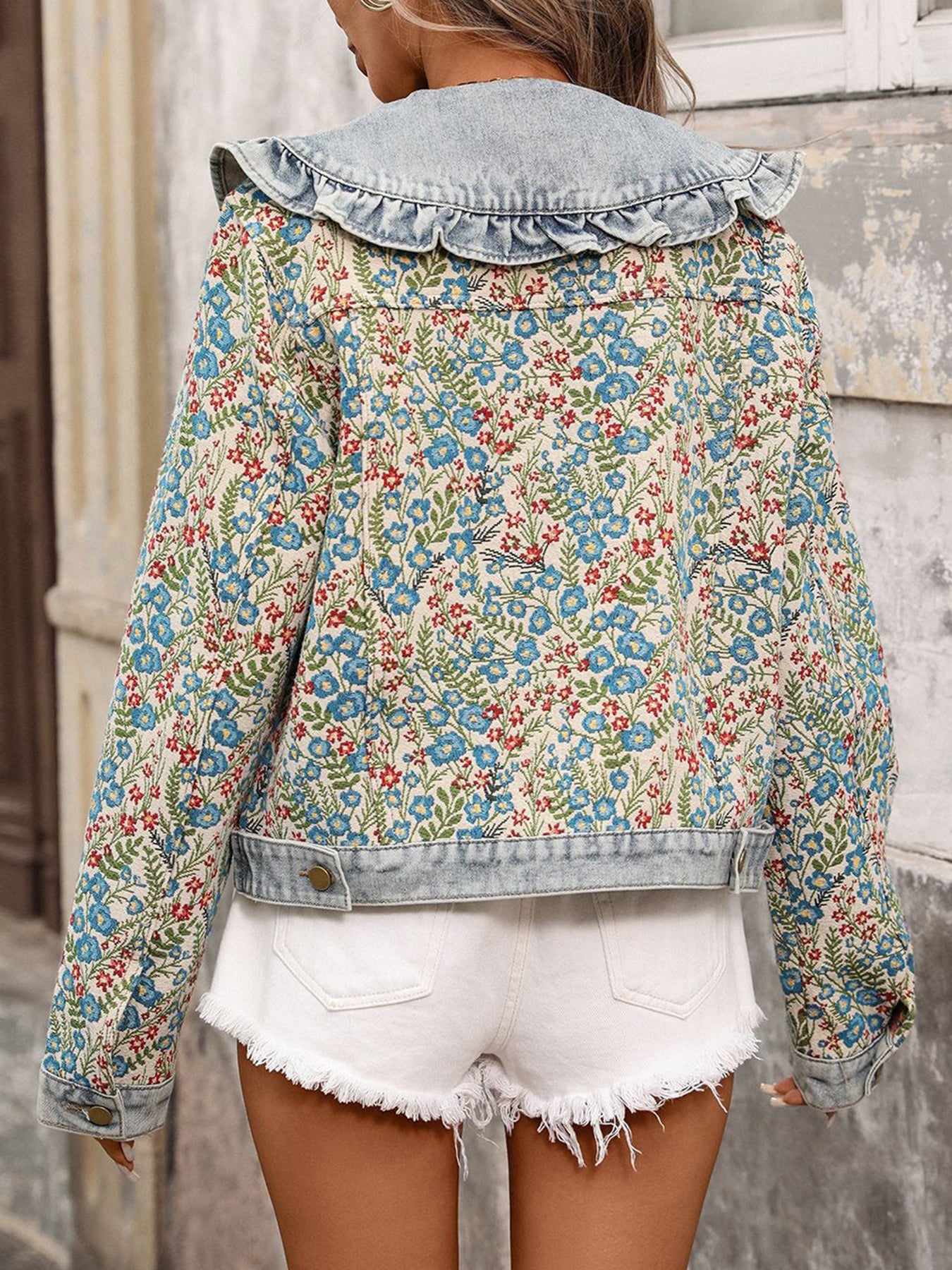 A woman shown from the back wearing a cropped denim jacket with a large ruffled collar and a blue, red, and green floral print, paired with white frayed denim shorts against a rustic stone wall.