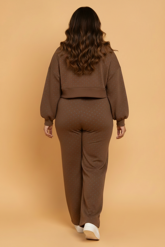A woman viewed from behind wearing a matching brown knit lounge set with a cropped long-sleeve top and wide-leg pants, standing against a solid warm beige studio background.