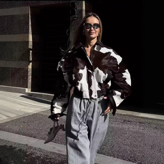 A woman wearing a black and white cow-print puff-sleeve jacket and light gray wide-leg sweatpants, standing outdoors in front of a building entrance.