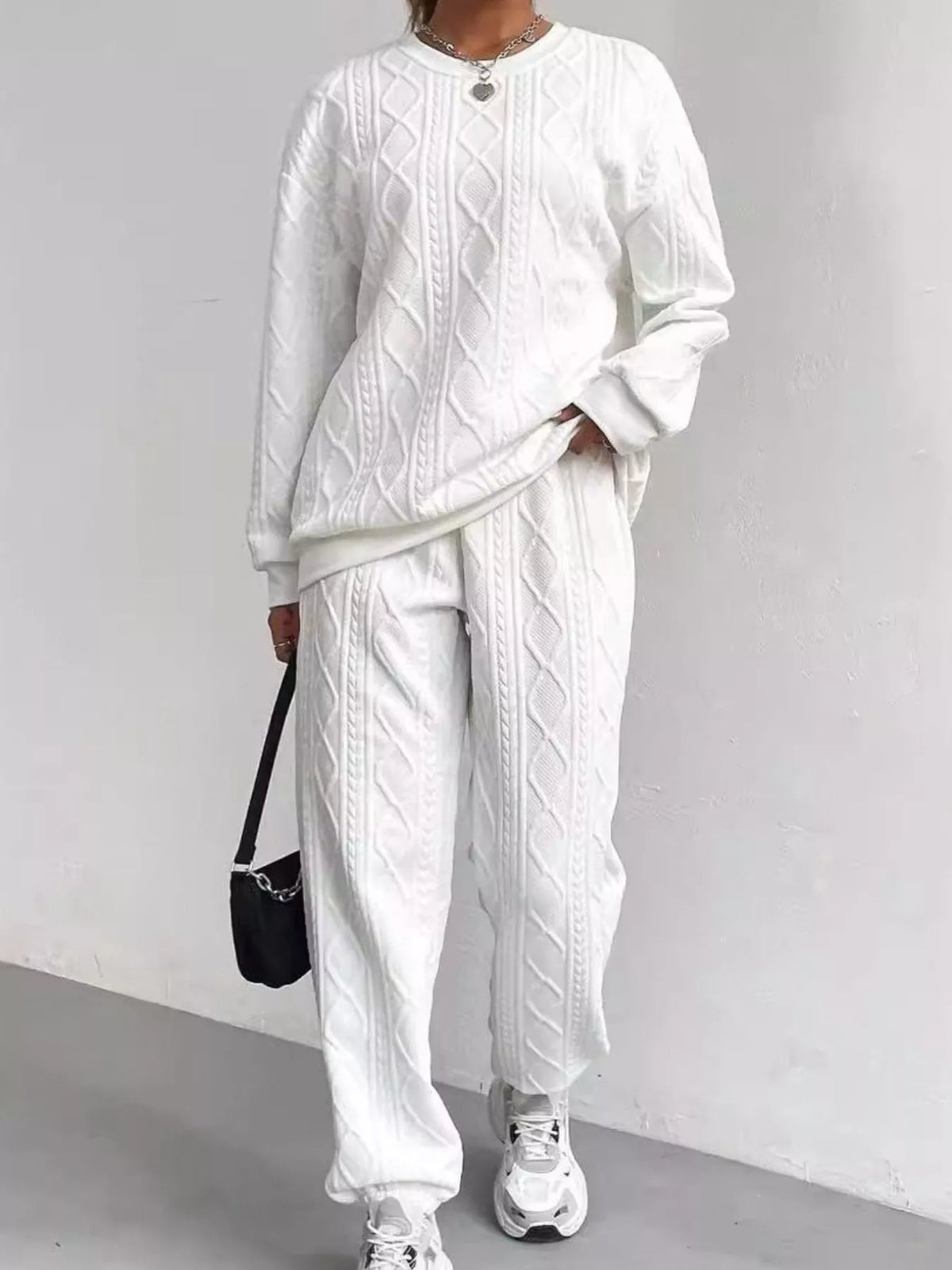 Person wearing a white cable knit sweater and pants with sneakers, holding a black bag against a plain background.