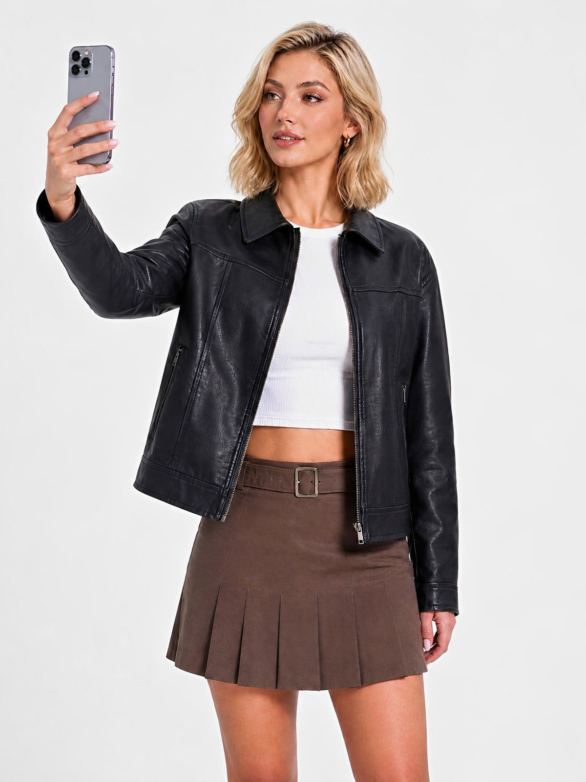 A woman modeling a black faux-leather jacket over a white crop top and brown pleated mini skirt, taking a mirror-style selfie against a light gray studio background.