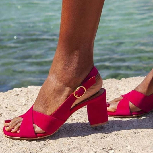 A close-up of a person wearing bright pink slingback high-heeled sandals with crisscross toes and a gold buckle, standing on a sandy surface with water in the background.