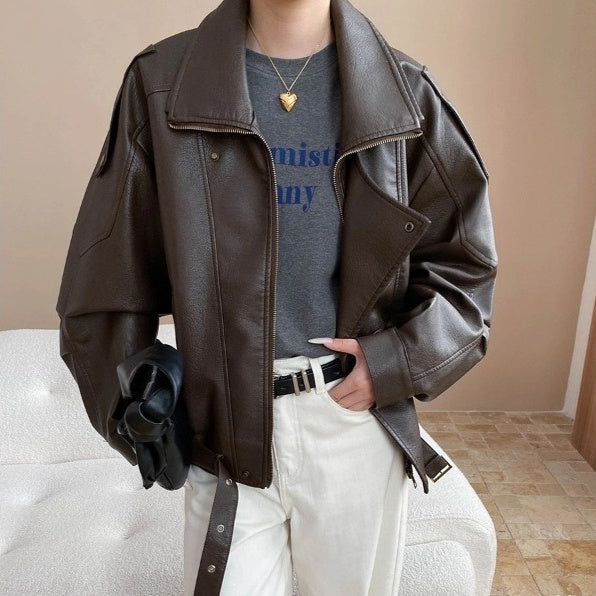 Person wearing a brown leather jacket over a gray shirt with text, white pants, and holding a black bag.