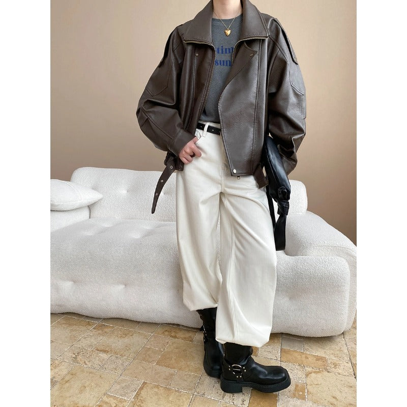 Person wearing a brown leather jacket and white pants standing in a room with a beige wall and white couch.
