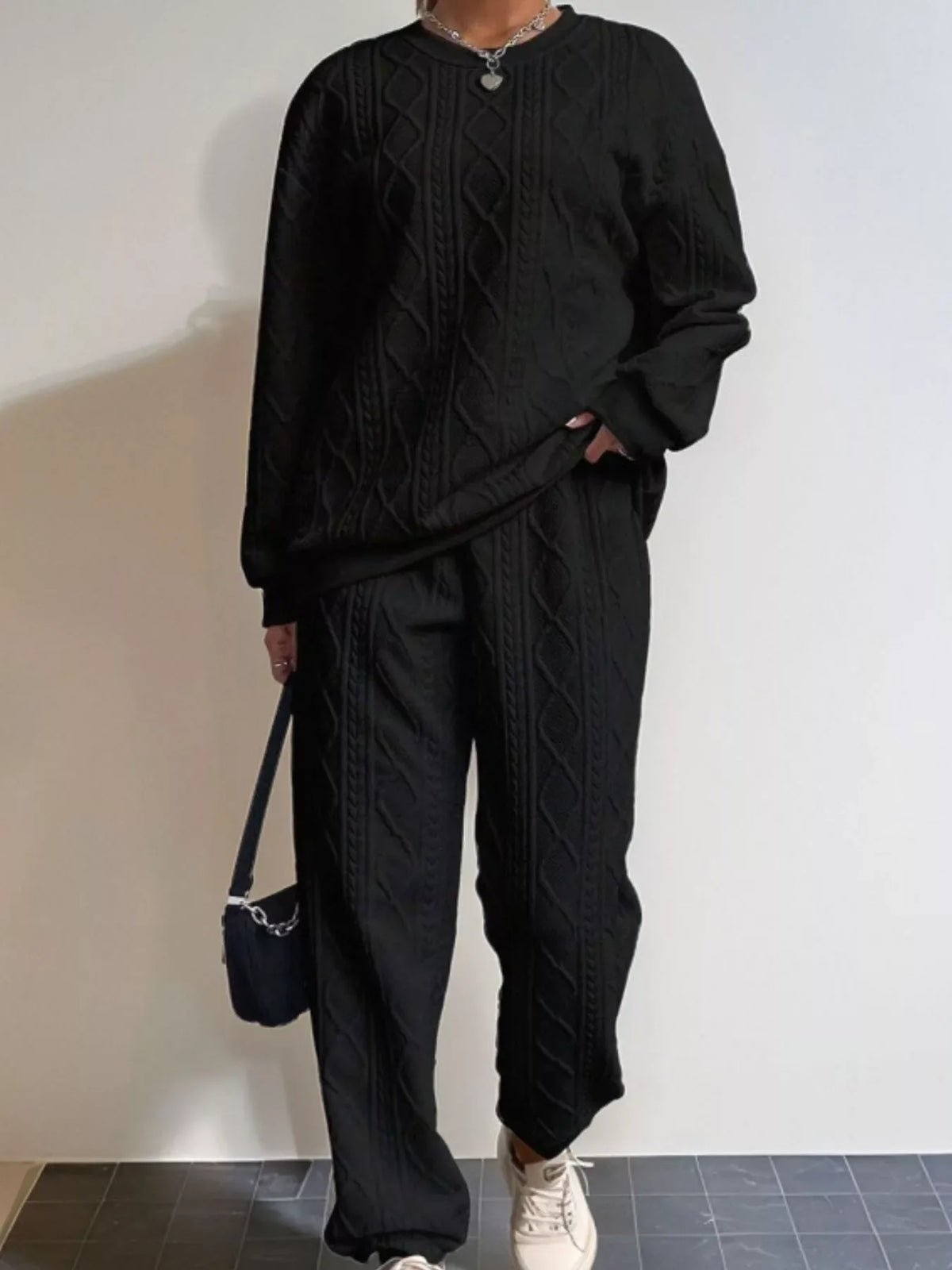 Person wearing a black cable knit sweater and pants with a white wall background