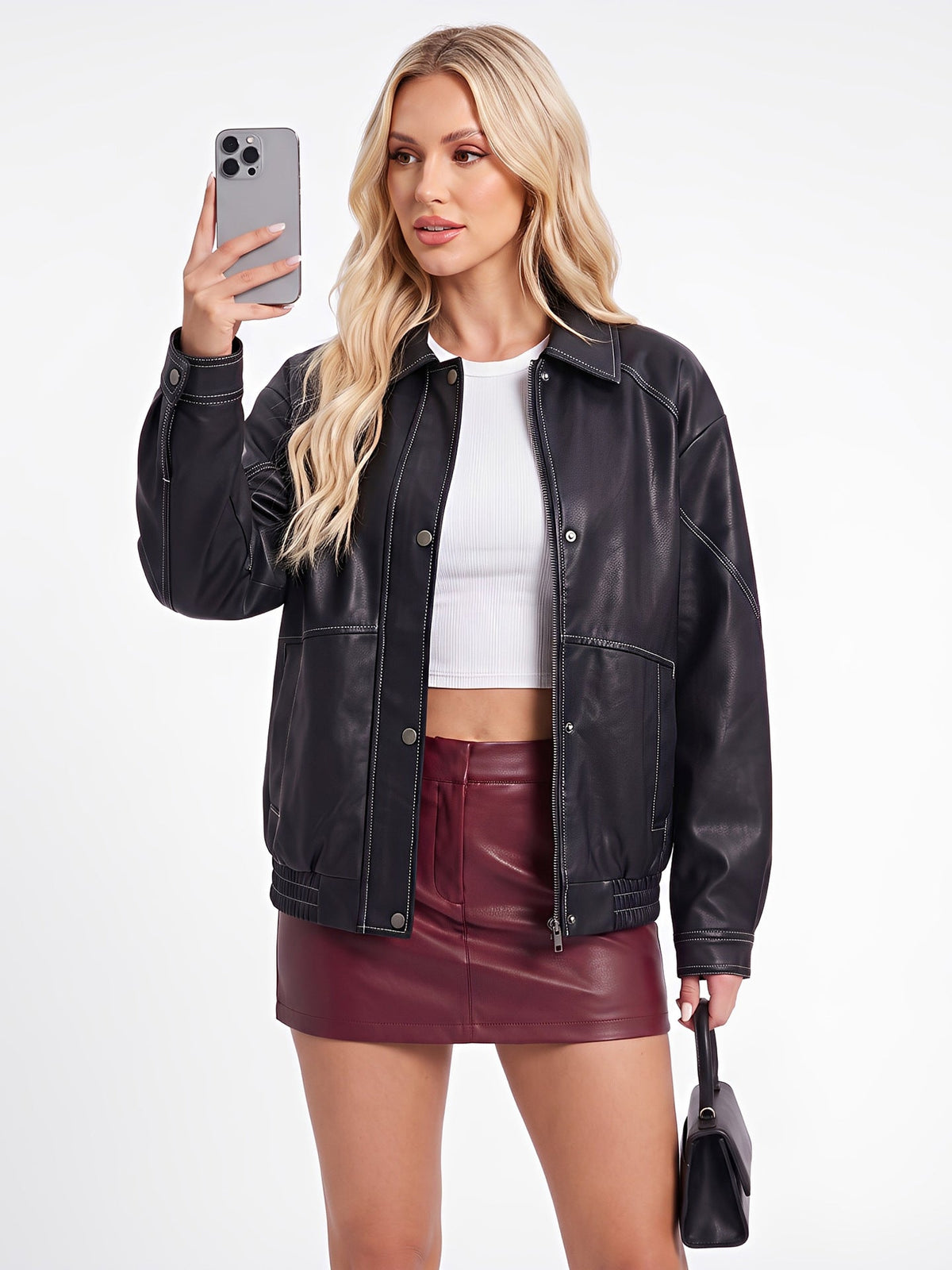 A woman modeling a black faux-leather jacket, styled with a white crop top, a burgundy faux-leather mini skirt, and holding a black handbag, while taking a mirror selfie with a smartphone against a plain white studio background.