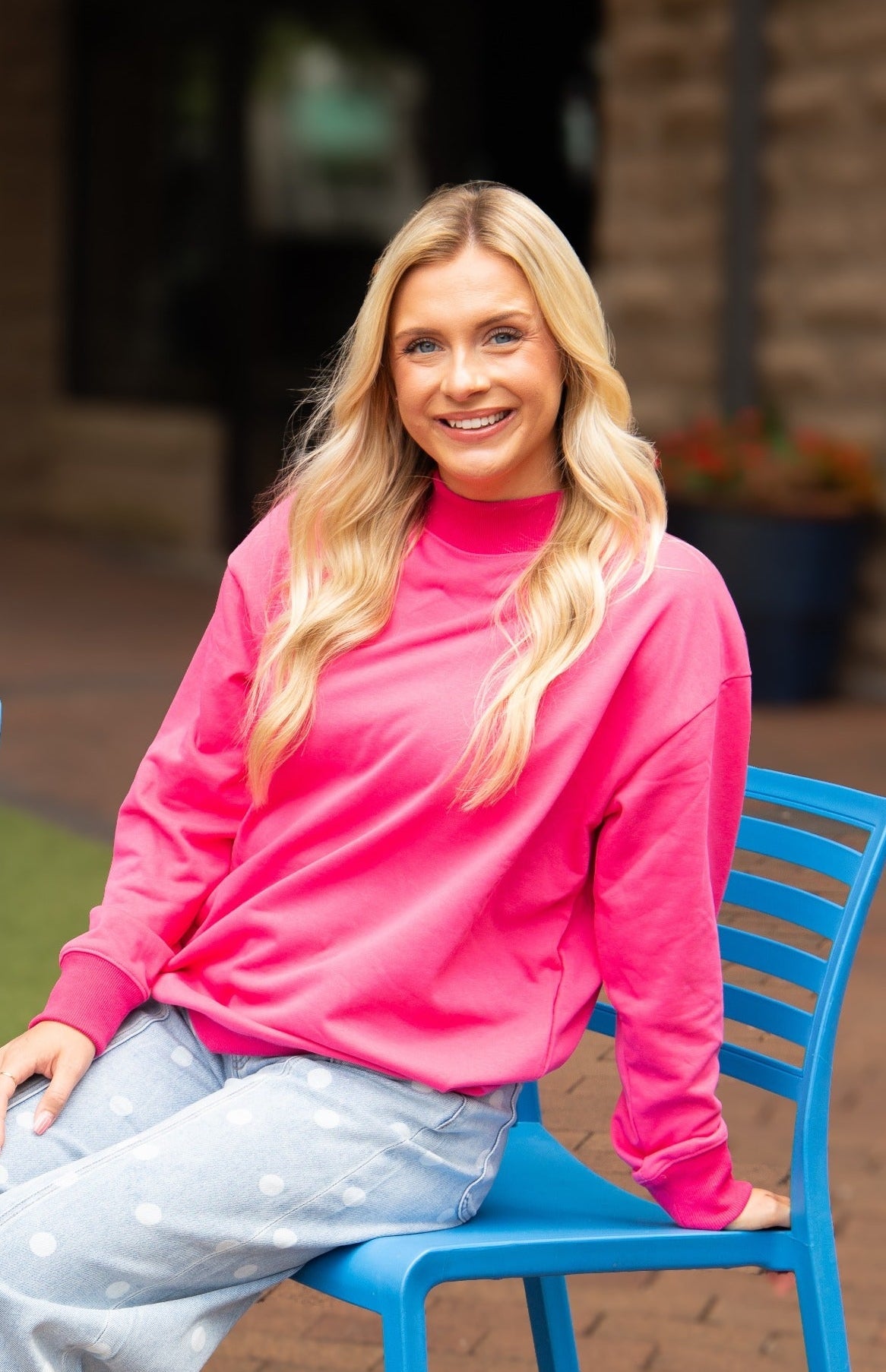 A smiling blonde woman wearing a bright pink mock-neck sweatshirt and light blue jeans, sitting on a blue chair outdoors.