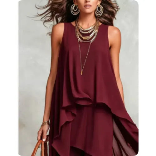 A woman wearing a sleeveless, deep burgundy dress with a layered, ruffled asymmetrical hem. She is accessorized with layered gold necklaces, large gold hoop earrings, and bracelets, holding a brown leather handbag against a textured gray background.
