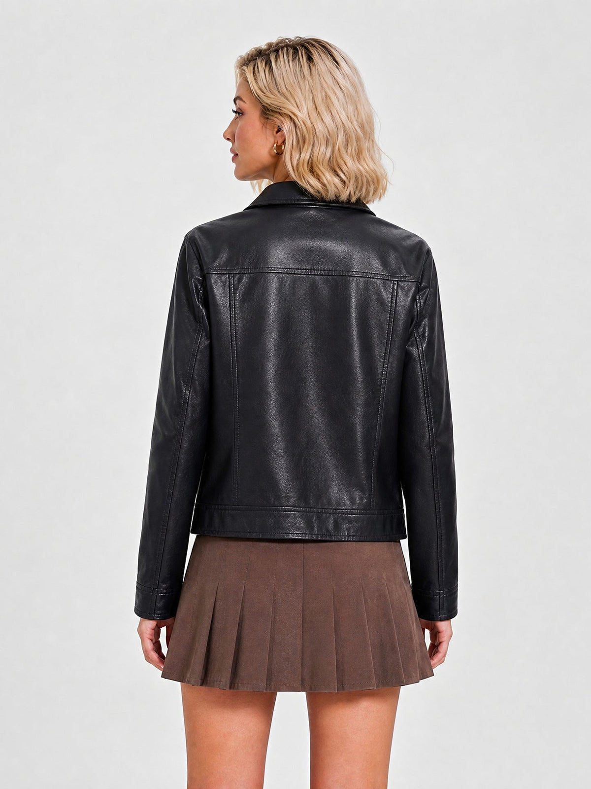 A woman shown from the back wearing a black faux-leather cropped jacket and a brown pleated mini skirt, photographed against a light gray studio background.