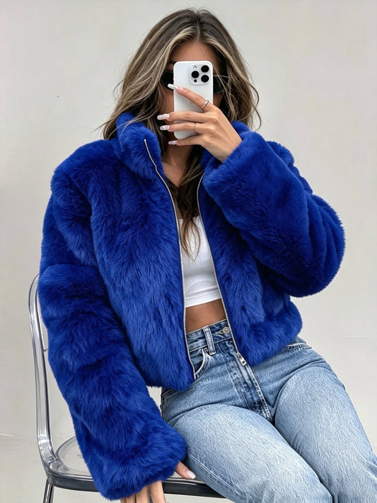 A woman wearing a bright royal blue faux fur jacket, white crop top, and light blue jeans, taking a mirror selfie while sitting on a clear chair against a plain light gray wall.