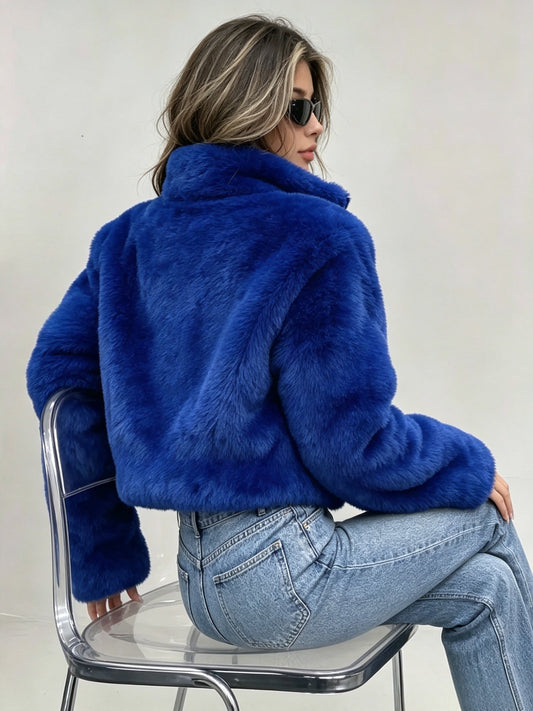 A woman wearing a bright royal blue faux fur jacket and light wash blue jeans, seated on a clear plastic chair against a plain off-white background.  Show the back of the Jacket.